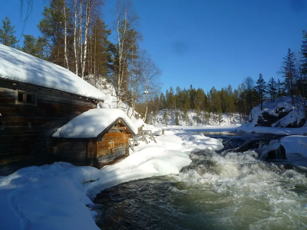 A winter photograph of Myllykoski in Finland, with snow on irregular banks beside dark moving water